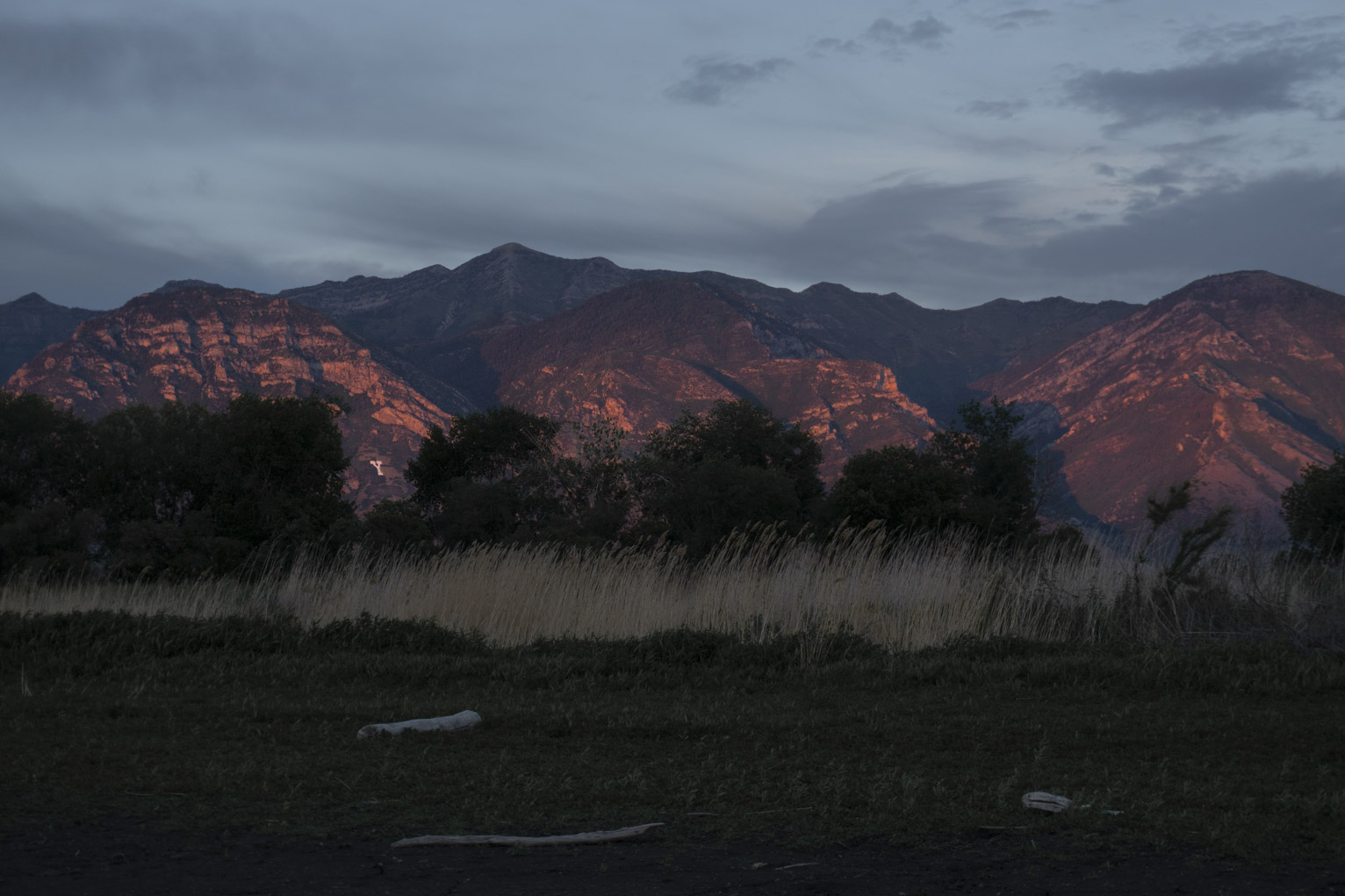 Behind the green grass, yellow grass, behind that, trees, and behind that sunset lit mountains with the university's white 'Y' painted on the mountain visible between trees